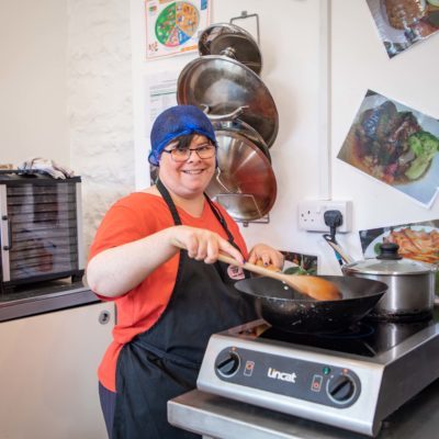 A lady wearing an apron is smiling as she stirs the dish she is cooking