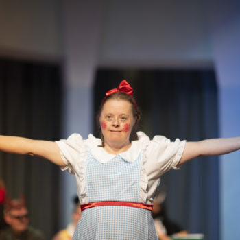A person we support wearing a costume in a play, wearing a blue checkered pinafore and red bow