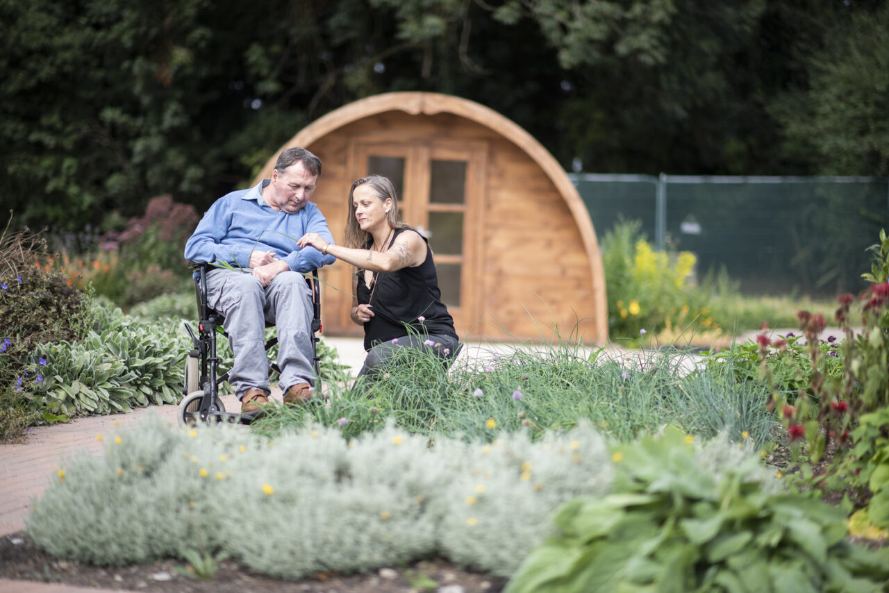 A person we support in a wheelchair and a staff member sitting in a garden together looking at a flower in Camphill Village Trust, UK charity