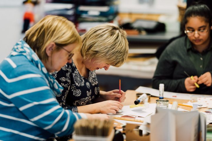 3 People sitting at a table in an arts and crafts workshop gluing something onto paper