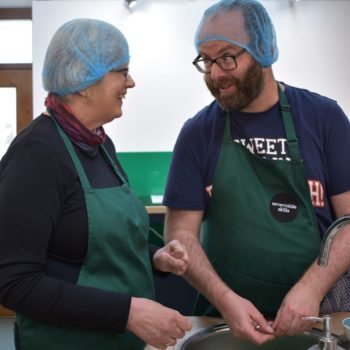 A person we support and staff member in the kitchen wearing aprons and hair nets preparing food and talking