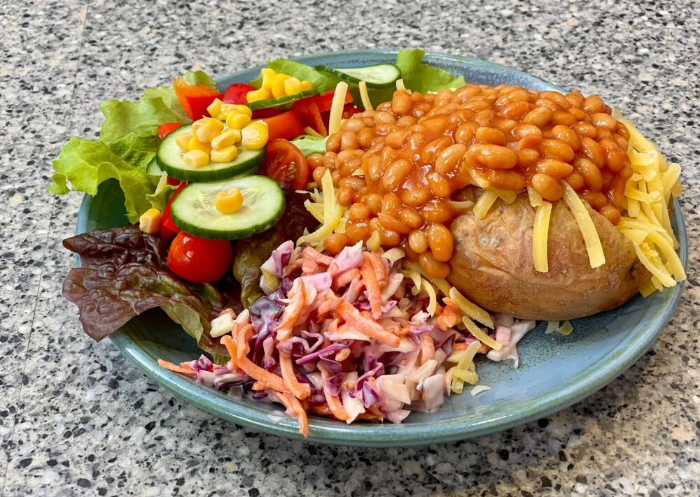 A slice of homemade quiche is served on a blue-green plate with a fresh side salad and chutney. The salad includes lettuce leaves, sliced cucumber, cherry tomatoes, diced red pepper, and a pink coleslaw-style slaw. A small white ramekin filled with chutney sits behind the quiche. The plate is presented on a wooden table, with the background softly blurred to keep the focus on the colourful lunch dish.