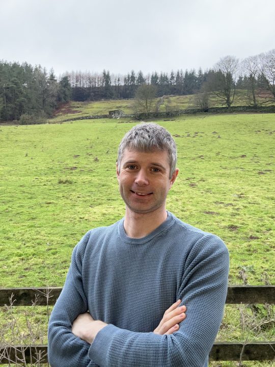 John leans against a fence in front of a field - his arms are crossed and he is smiling