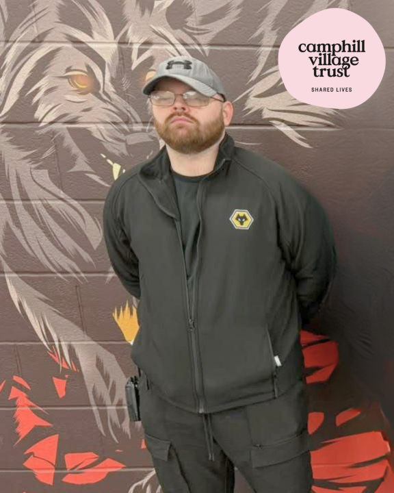 Shane is standing in front of a colourful wall featuring a Wolves mural. The Camphill Village Trust Shared Lives logo (pink circle) appears top right.