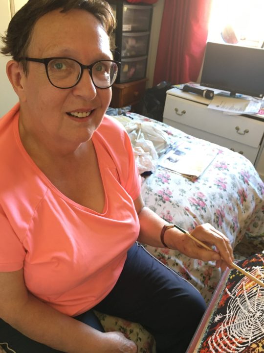Christina sitting painting, she is wearing an orange top and black glasses. She is looking up at the camera.