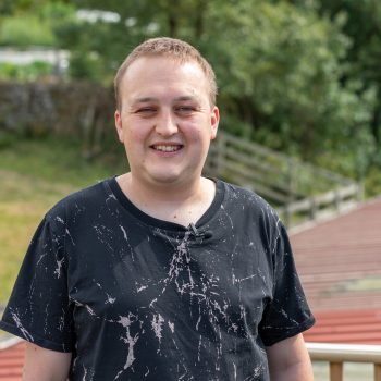 Simon is facing the camera smiling, in front of a natural background.