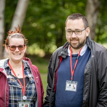 Nicki and Ed stand in front of a natural background. They are smiling at the camera, wearing coats and their Life of Opportunity Reviewer lanyards.