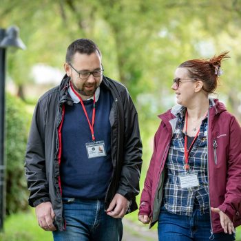 Nicki and Ed are walking outside talking and laughing together. Both are wearing their Life of Opportunity Reviewer lanyards.