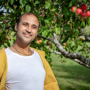 Deepak is standing by an apple tree, he is smiling wearing a yellow hoodie and a white shirt.
