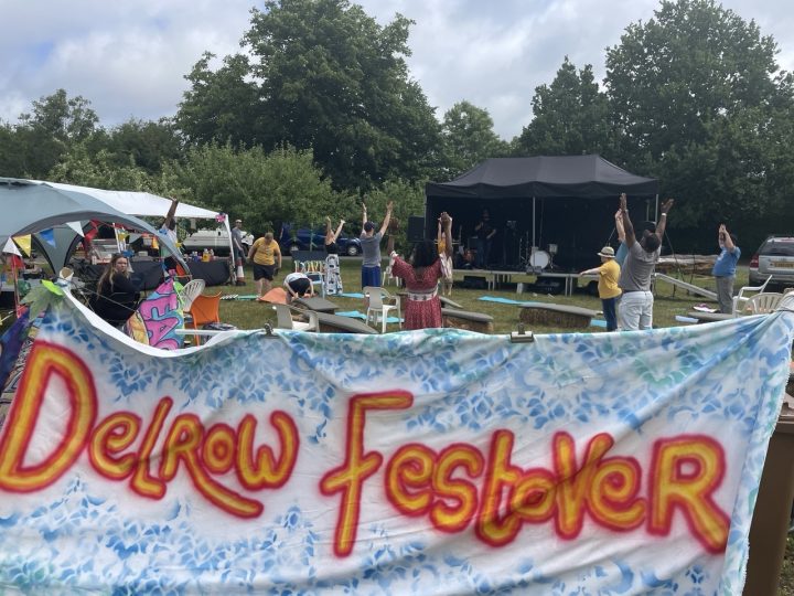 A photo of a stage outside, in the foreground is a spray painted sign saying "Delrow Festover", there are people having fun, doing stretches in front of the stage, 