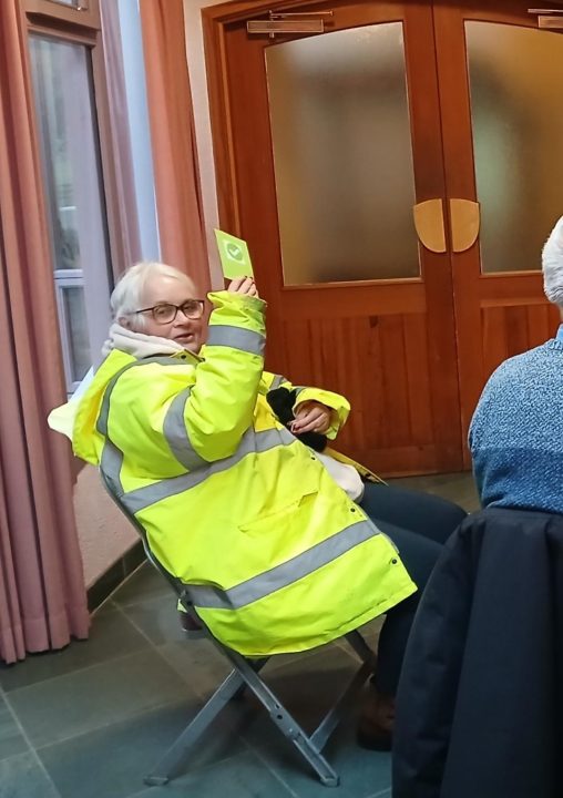 A member of our Botton community is sitting in a chair holding up a green card as part of an activity, she is wearing a high vis jacket.