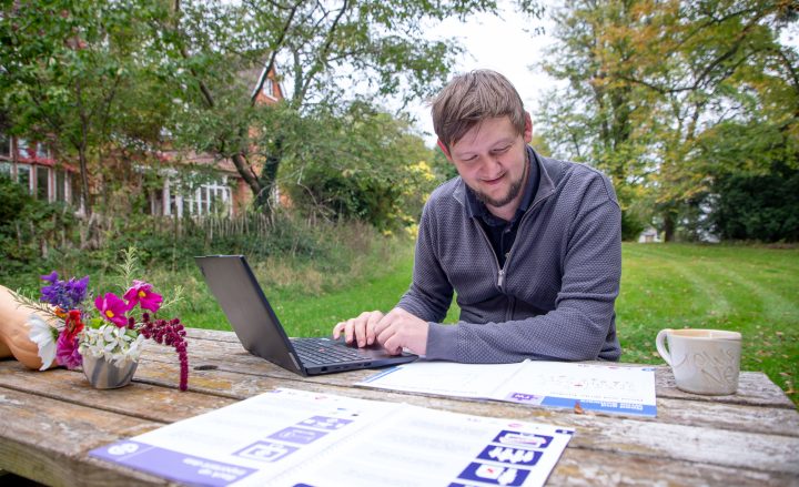 A man sitting outside at a picnic bench with a laptop, he is looking down at easy read digital inclusion guides.