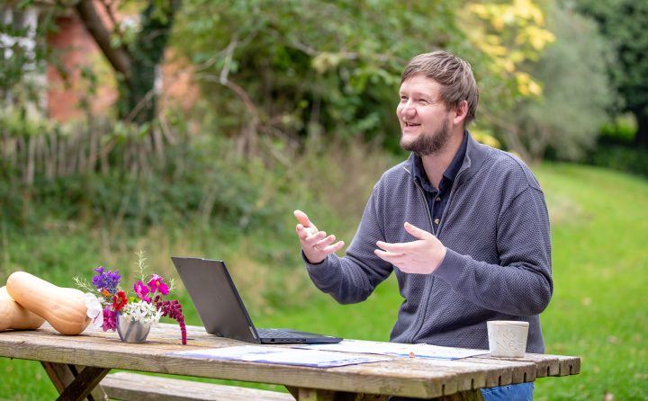 Alex is sitting at a picnic bench outside with a laptop. He is looking away from the camera and seems to be explaining something, using his hands.