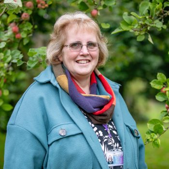 Photo of Miriam Goldby, she is standing near an apple tree and smiling at the camera.