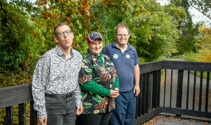 Three community members standing on wooden decking in front of trees