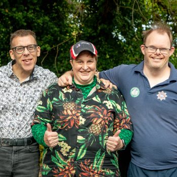 Three members of the Digital Inclusion group are standing outside with their arms around each other, smiling. The person in the middle has their thumbs up.