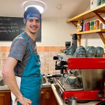 A person we support helping in a cafe kitchen, he is wearing a hair net and smiling.