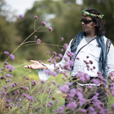  A man wearing bohemian clothing and sunglasses stands peacefully in a wildflower meadow.