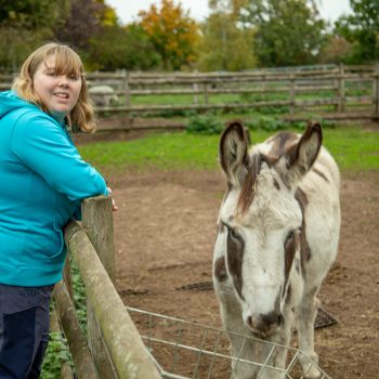 A woman in a blue goat leans on the fence of a donkey's pen