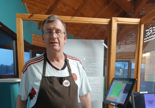 A man in a Camphill Village Trust apron behind a till