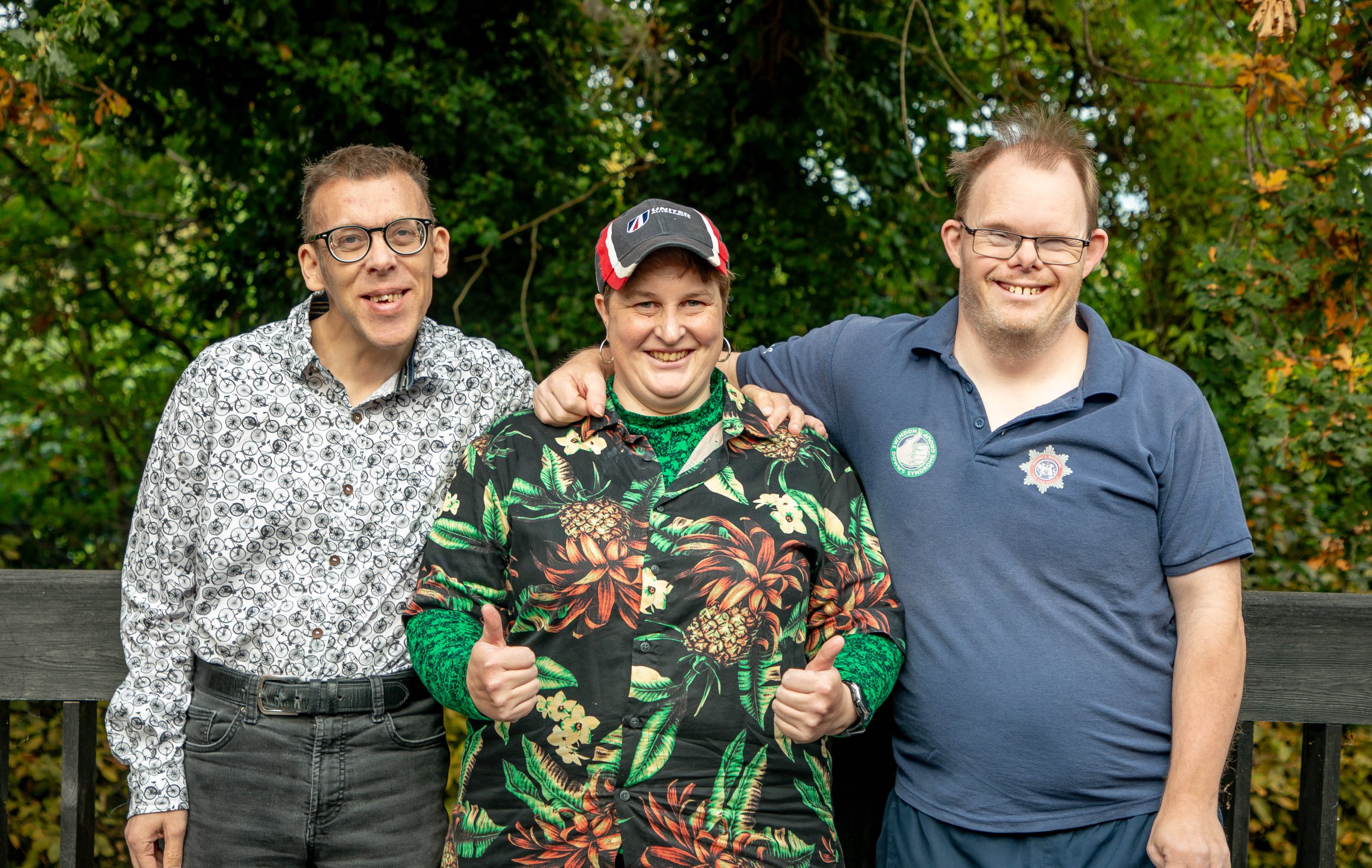 Three people we support stand with their arms around each other smiling. The person in the middle has their thumbs up.