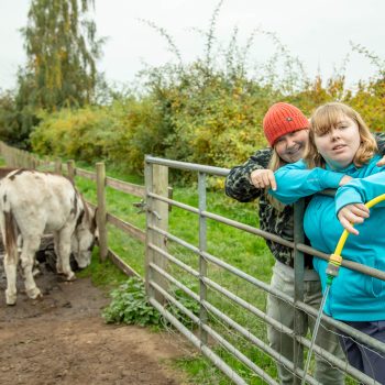 A person we support is leaning on a fence, filling a bucket with water from a hose. A support worker has her arm around her, smiling. There are donkeys in the field beyond the fence.