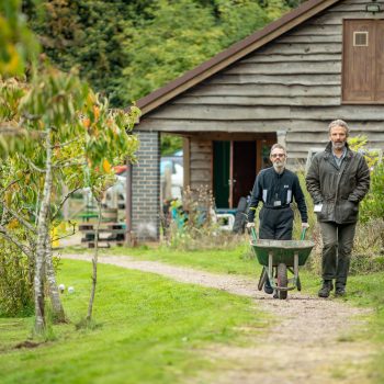 A support worker walks alongside a person we support along a path in an orchard. the person we support is pushing a wheelbarrow.