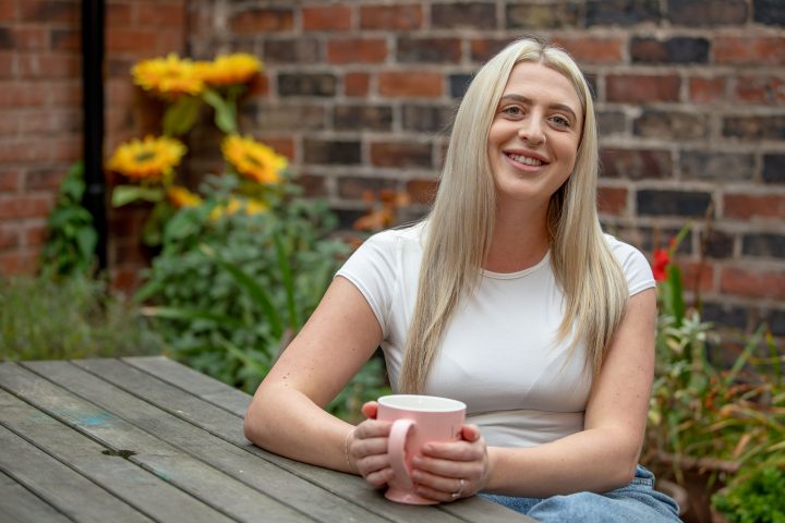Hollie, Stourbridge Registered Manager, sits at a picnic table, holding a pink mug, there are yellow sunflowers in the background.