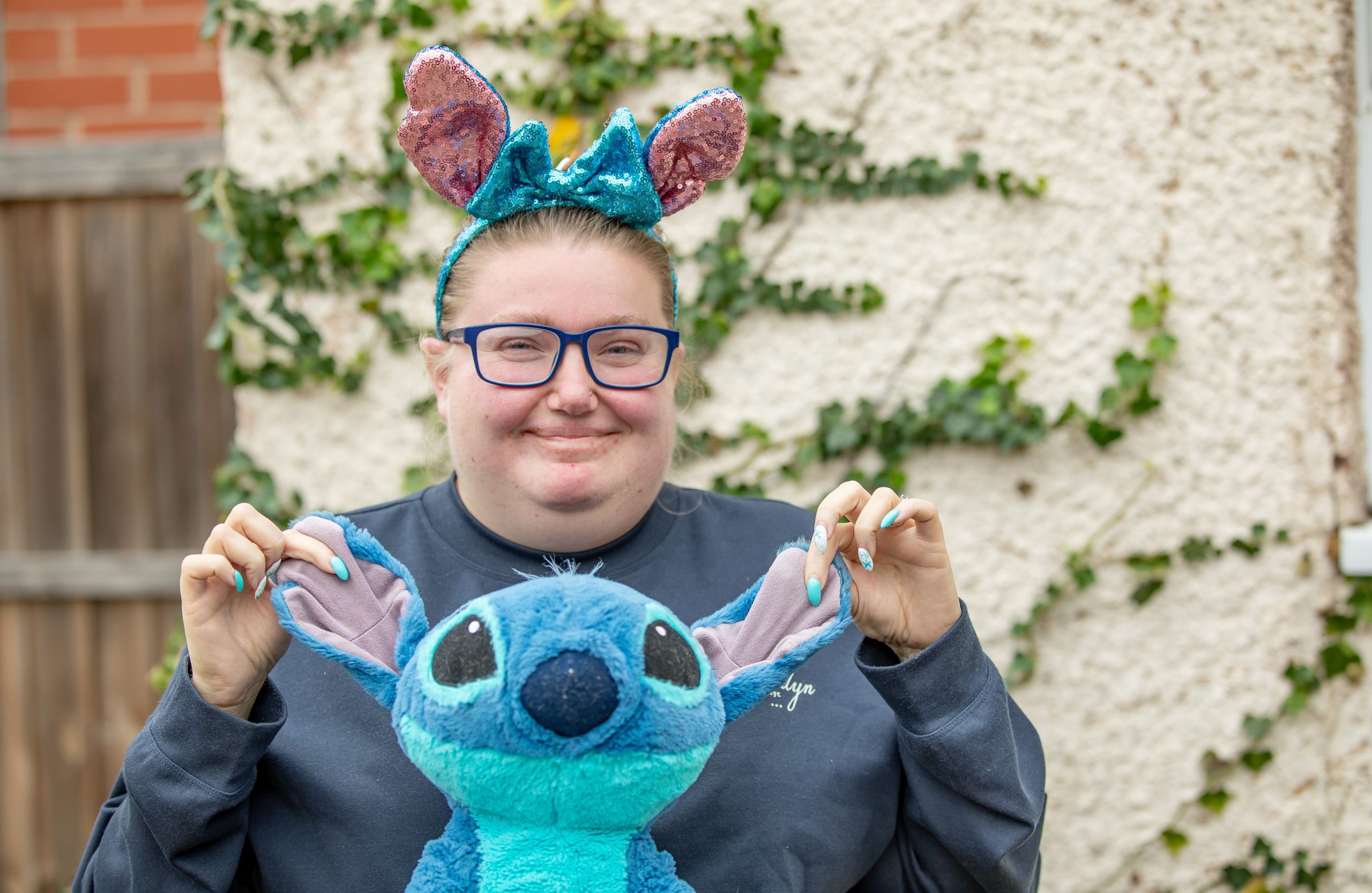 Sophie is standing in her back garden in front of an ivy-covered wall. She is smiling and wearing sparkly blue and pink "Stitch" ears from the film Lilo and Stitch. She is holding up a stitch character by the ears and showing it to the camera.