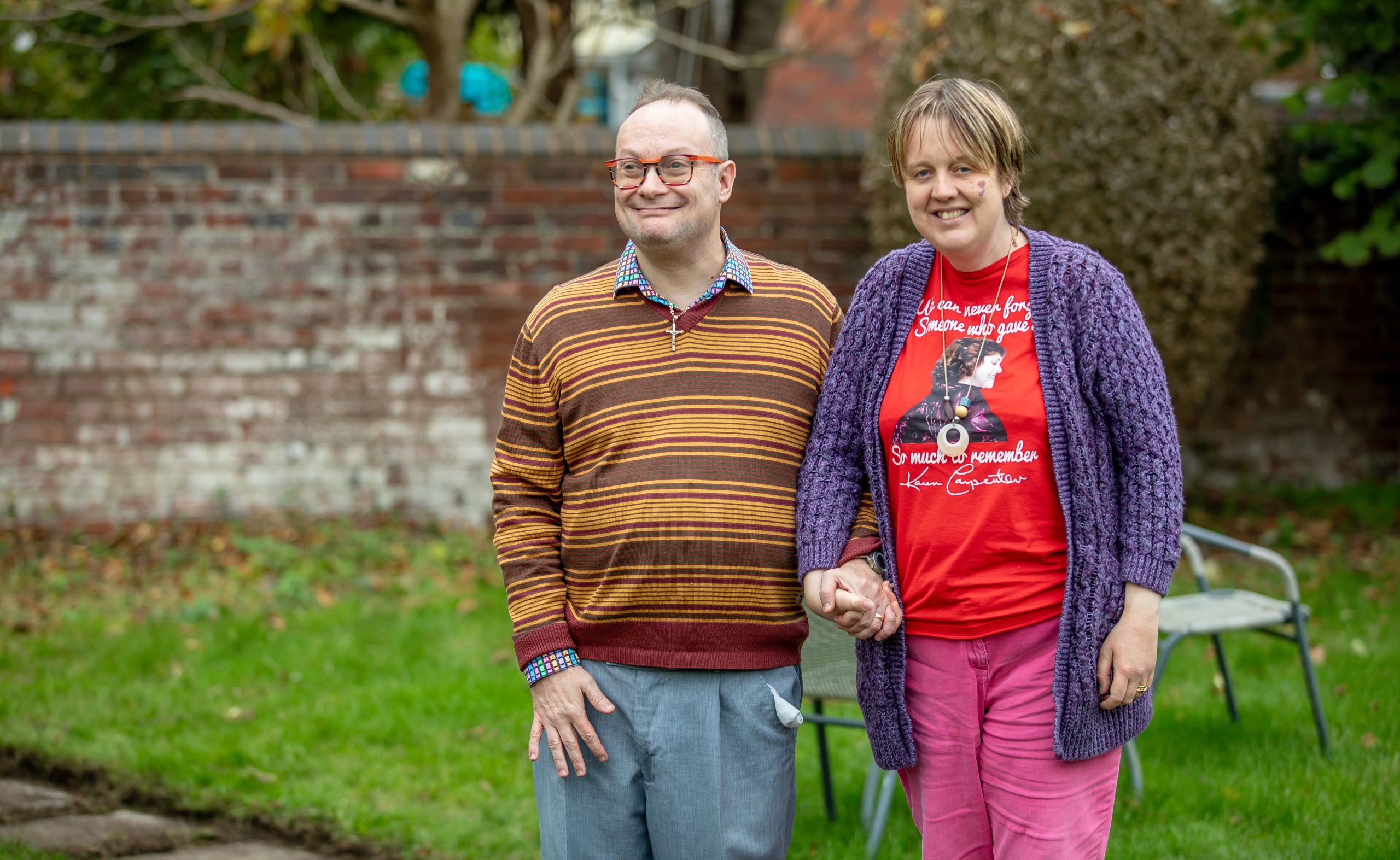 Andrew and Karen stand in their garden in the autumn. They are holding hands tightly and smiling.