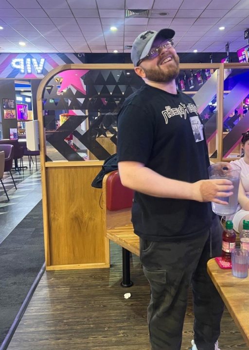 A man in a peaked hat is standing holding a pitcher in the middle of a bowling arcade, he is smiling.