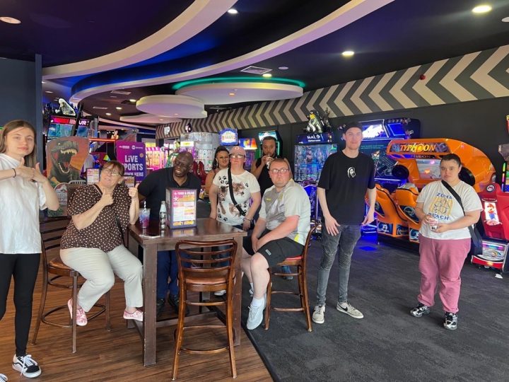 A group of people stand around a table in a bowling arcade - lots of them have their thumbs up
