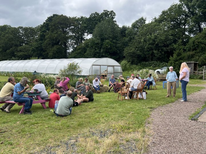 A group of people sit and chat outdoors on picnic benches, chairs and hay bales beside a large polytunnel and flower garden. The gathering takes place on a grassy area surrounded by trees, with a gravel path running alongside the group.