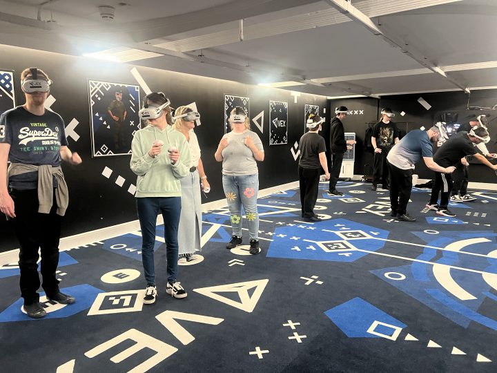 A group of people wearing virtual reality headsets take part in an immersive gaming experience inside a spacious indoor activity room. The room features black walls with geometric designs and a blue patterned floor, while participants use handheld controllers to interact with the virtual environment.