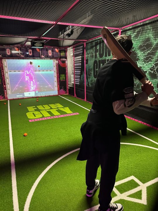 A person stands in an indoor interactive batting cage holding a cricket bat, facing a large digital screen displaying a virtual bowler. The activity area has artificial turf, neon pink framing and Fun City branding, with several orange balls scattered on the floor.