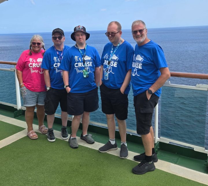 Five people stand together on the outdoor deck of a cruise ship with the sea stretching into the background. Four are wearing matching blue “It’s Cruise Time” T-shirts and one is wearing a pink version. The group poses beside the ship’s glass railing on a bright day with calm ocean views behind them.