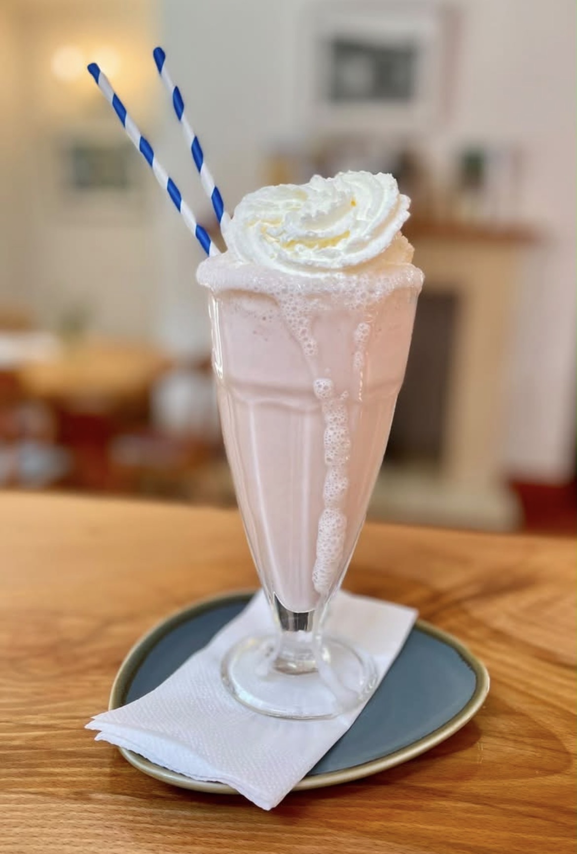 A tall strawberry milkshake topped with whipped cream is served in a classic glass on a blue-grey saucer with a folded white napkin. Two blue-and-white striped paper straws sit in the milkshake, which has foam overflowing slightly down the side of the glass. The drink is placed on a wooden table, with the bright café interior softly blurred in the background.