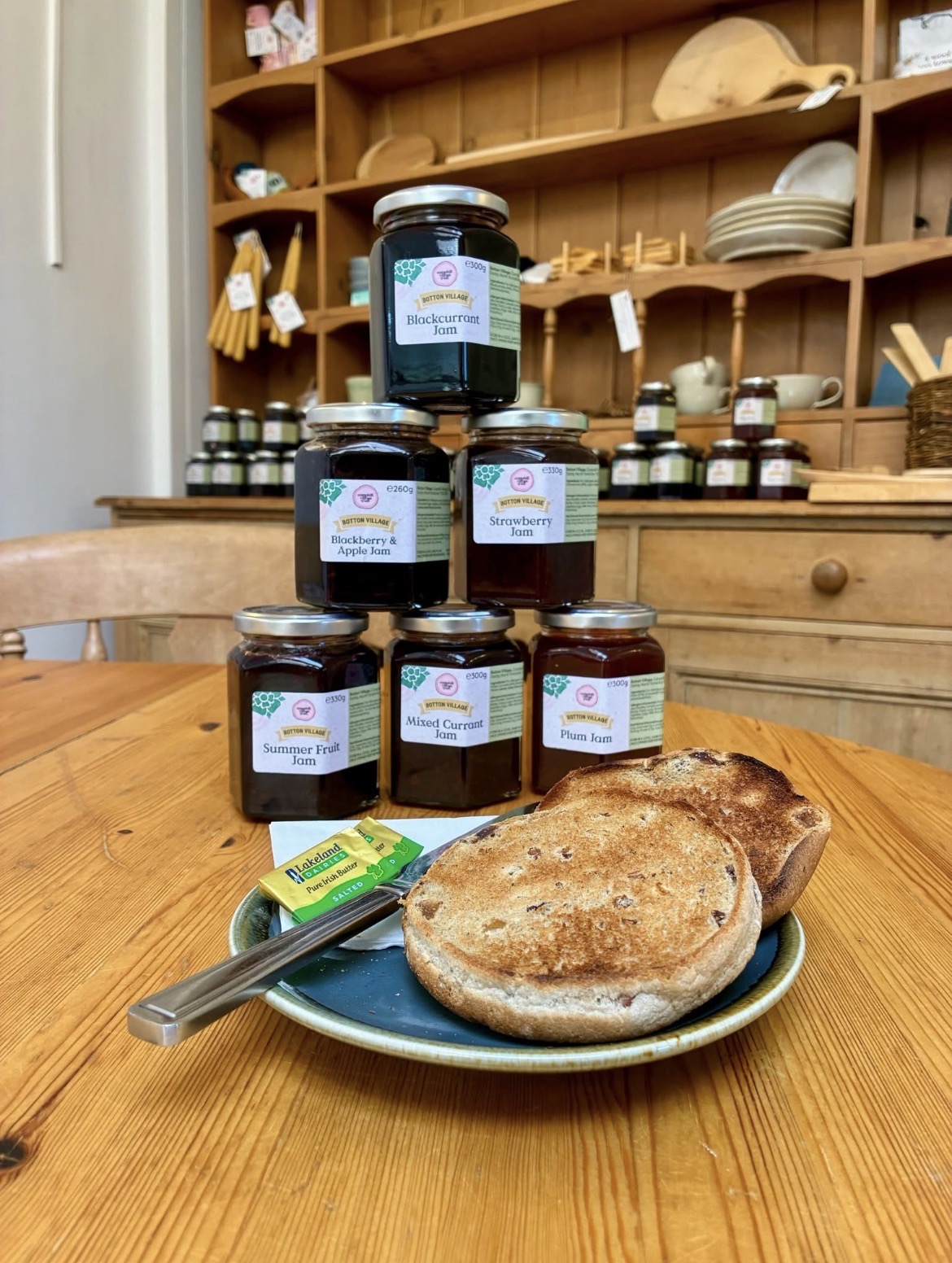 A plate of toasted fruit bread with a butter portion and knife sits on a wooden table in a café. Behind it, six jars of Botton Village jam are stacked in a pyramid, including blackcurrant, strawberry, plum, mixed currant, summer fruit, and blackberry & apple varieties. Wooden shelves displaying cheeses, crockery, and additional jars create a warm, rustic café setting in the background.