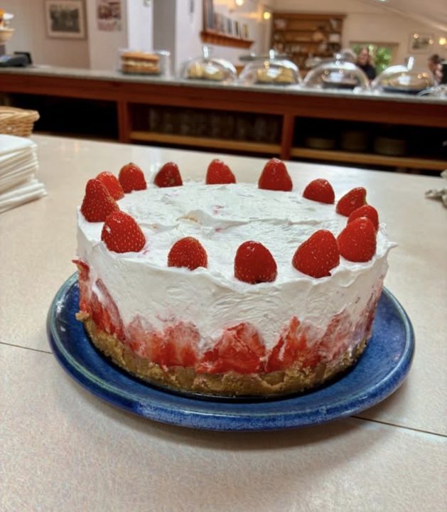A homemade strawberry cheesecake sits on a blue plate on a café counter. The dessert features a biscuit base topped with a creamy filling and fresh strawberries visible around the sides, finished with a layer of whipped cream and whole strawberries arranged around the top. In the background, covered cake stands and shelves are softly out of focus, creating a warm and welcoming café setting.