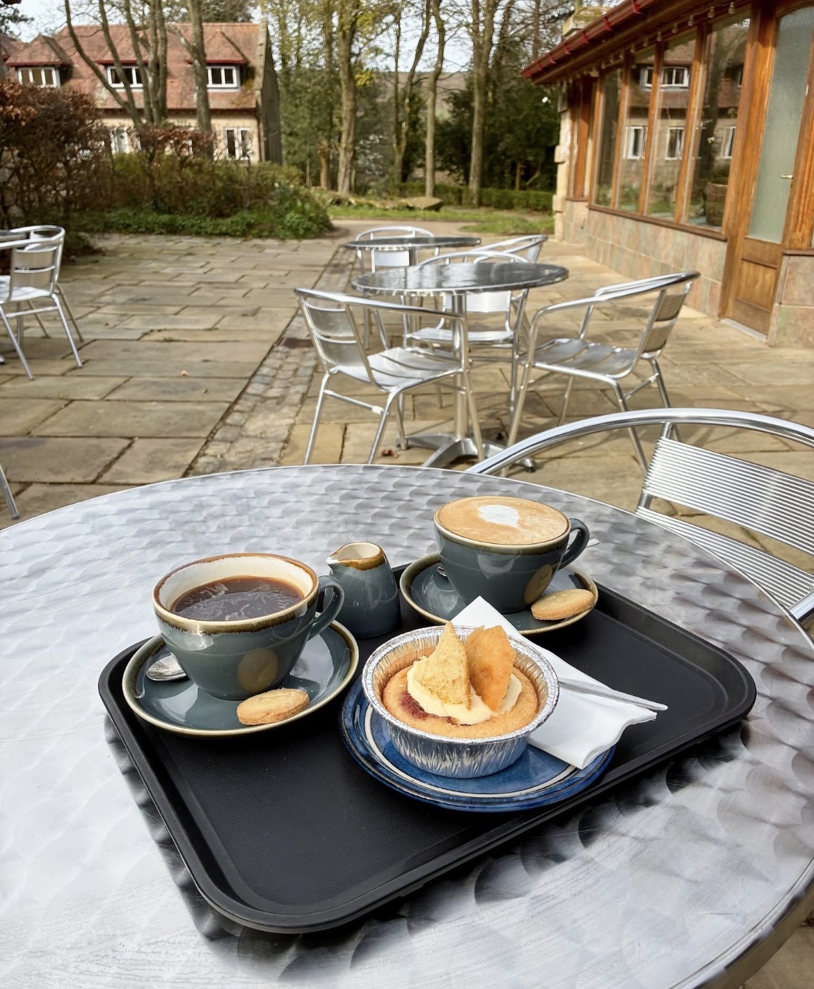 A tray with two hot drinks and a freshly baked scone sits on a metal café table on an outdoor patio. One cup contains tea and the other a frothy coffee, accompanied by a small jug of milk and biscuits. The scone is served with cream and jam on a blue plate. In the background, silver café tables and chairs are arranged on a stone terrace beside a building with large windows, with trees and gardens creating a peaceful countryside setting.