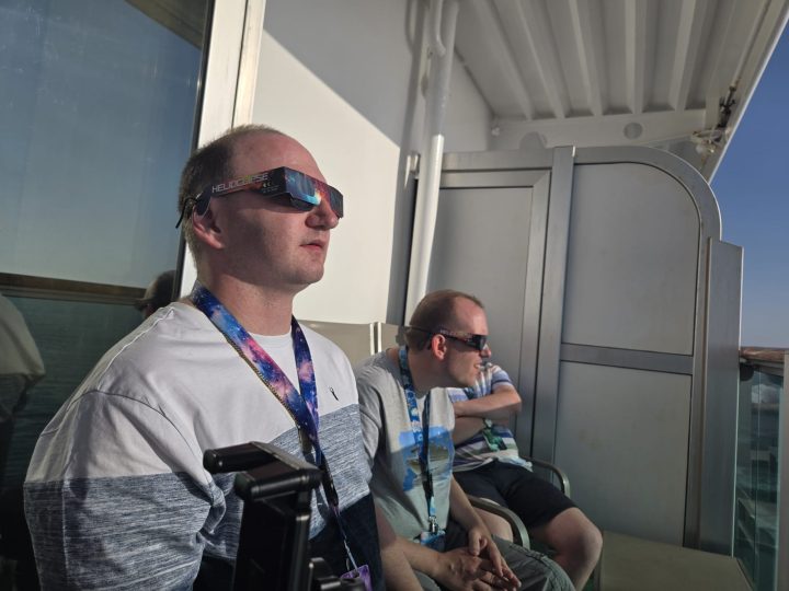 Three people sit on a cruise ship balcony wearing eclipse-viewing glasses while looking toward the sky. The foreground shows one person seated near the balcony door, with two others sitting further back. Bright sunlight illuminates the balcony, with the sea visible beyond the railing.