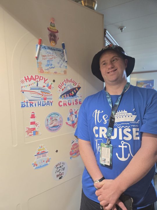 A person wearing a blue “It’s Cruise Time” T-shirt, a bucket hat and a lanyard stands beside a cabin door decorated with cruise-themed birthday decorations. The door features signs reading “Happy Birthday Cruise,” pictures of cruise ships, a birthday cake, a lighthouse and a personalised birthday message board with the name John. The scene appears to be aboard a cruise ship.