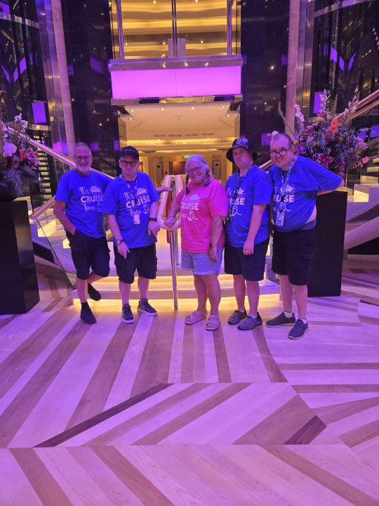 Five people wearing matching cruise-themed T-shirts pose together in the brightly lit central atrium of a cruise ship. Four wear blue “It’s Cruise Time” T-shirts and one wears a pink version. The group stands between two floral displays in front of a staircase and glass lift, with purple and gold lighting throughout the ship’s interior.