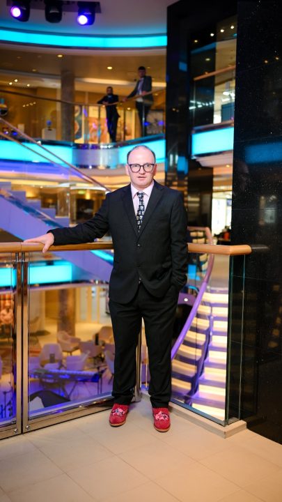 A person wearing a dark suit, patterned tie and glasses stands beside a glass balcony railing in a cruise ship atrium. The multi-storey interior is illuminated with blue and purple lighting, with staircases, seating areas and upper-level walkways visible in the background.