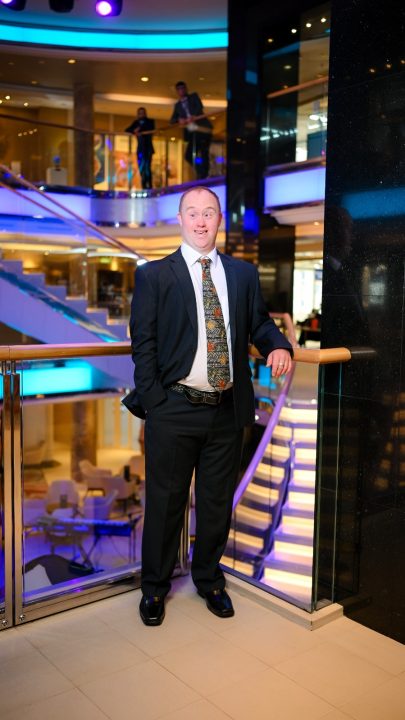 A person wearing a dark suit, patterned tie and glasses stands beside a glass balcony railing in a cruise ship atrium. The multi-storey interior is illuminated with blue and purple lighting, with staircases, seating areas and upper-level walkways visible in the background.