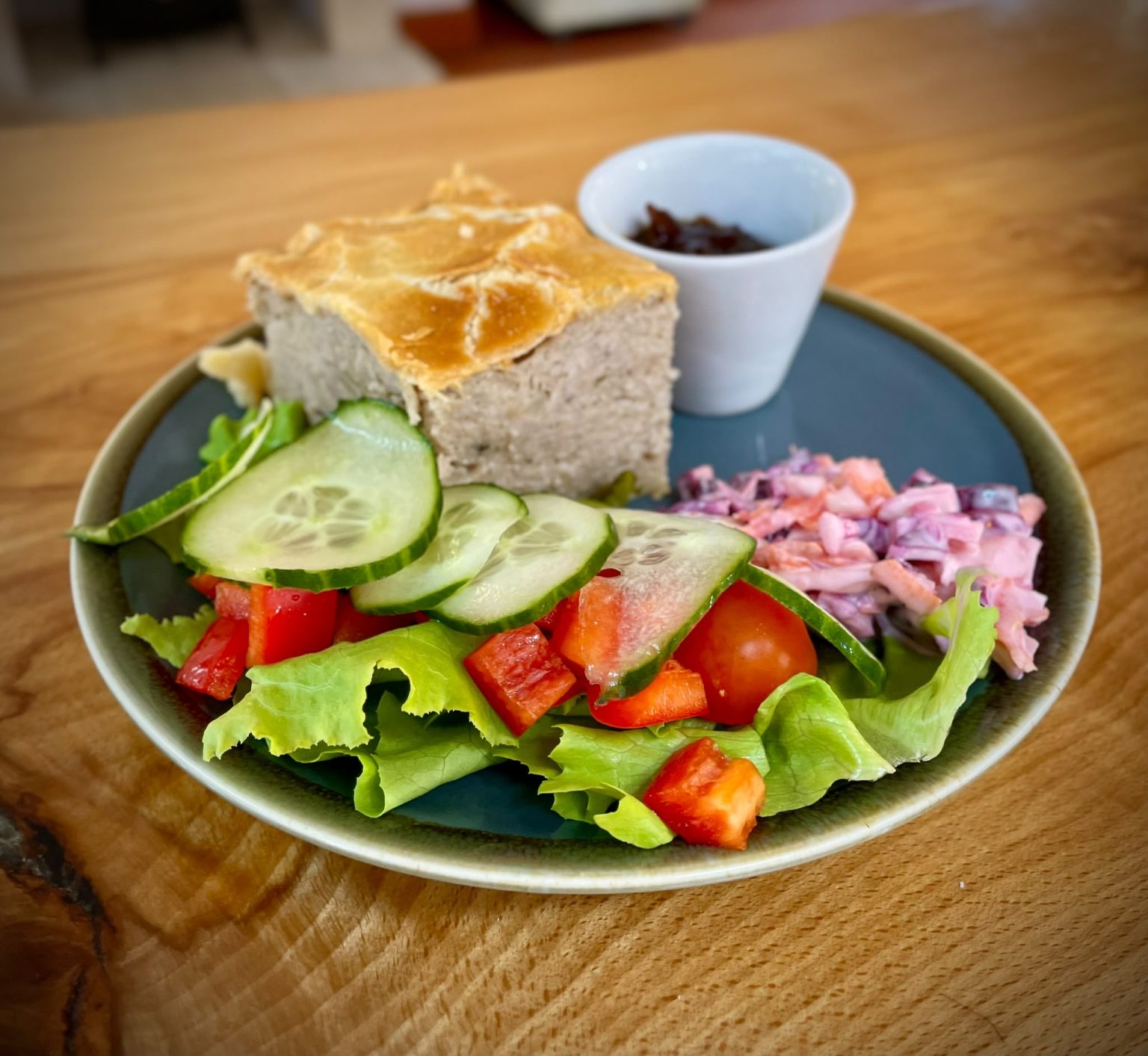 A slice of homemade quiche is served on a blue-green plate with a fresh side salad and chutney. The salad includes lettuce leaves, sliced cucumber, cherry tomatoes, diced red pepper, and a pink coleslaw-style slaw. A small white ramekin filled with chutney sits behind the quiche. The plate is presented on a wooden table, with the background softly blurred to keep the focus on the colourful lunch dish.