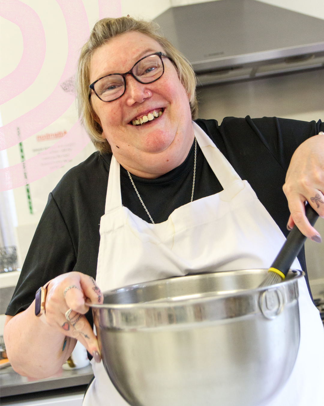 A person wearing a white apron stands in a kitchen holding a large metal mixing bowl and whisk. The kitchen includes a cooker and extractor hood in the background. The person is wearing glasses and several pieces of jewellery, and is preparing food at a work surface in a bright, modern kitchen.