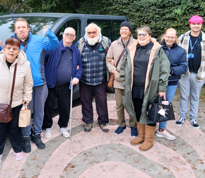 A group of eight adults stand together outdoors in front of a minibus and a hedge-lined background. Several people are wearing coats and jackets, and one person is using a walking stick. The group is gathered closely together on a paved area with a decorative pink pattern overlaid at the bottom of the image. The photo appears to have been taken during a group outing or trip.